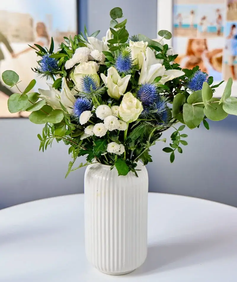 A bouquet of white and blue flowers with lush green foliage arranged in a tall white ribbed vase on a round table. A bouquet of white and blue flowers with lush green foliage arranged in a tall white ribbed vase on a round table.